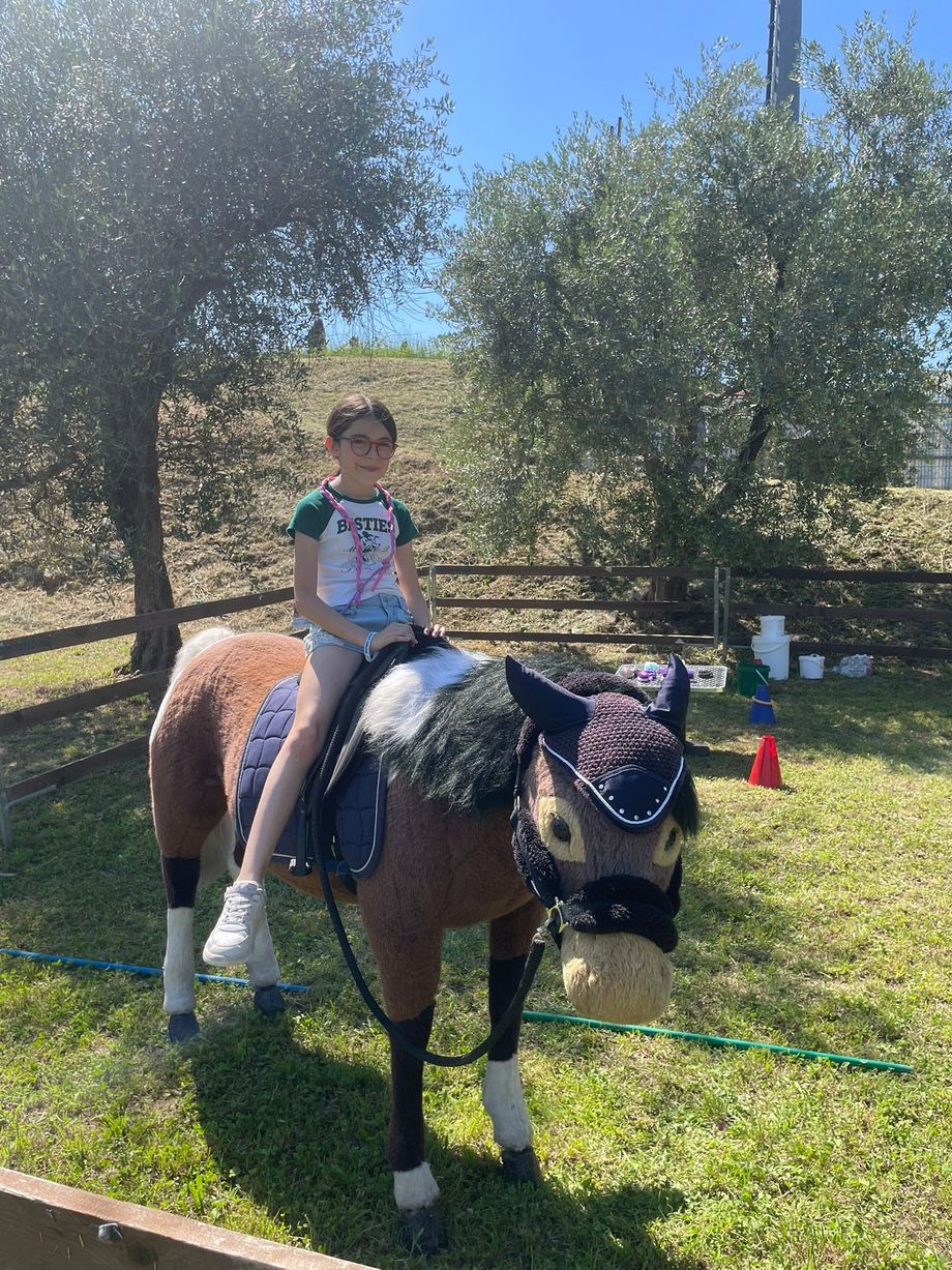 Una bambina sorride, seduta su un pony pezzato sellato, sotto un grande ulivo